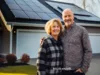 Middle aged couple standing on front of a house with solar panels on the roof. They smiling and looking at camera. Innovative energy systems to save your money.